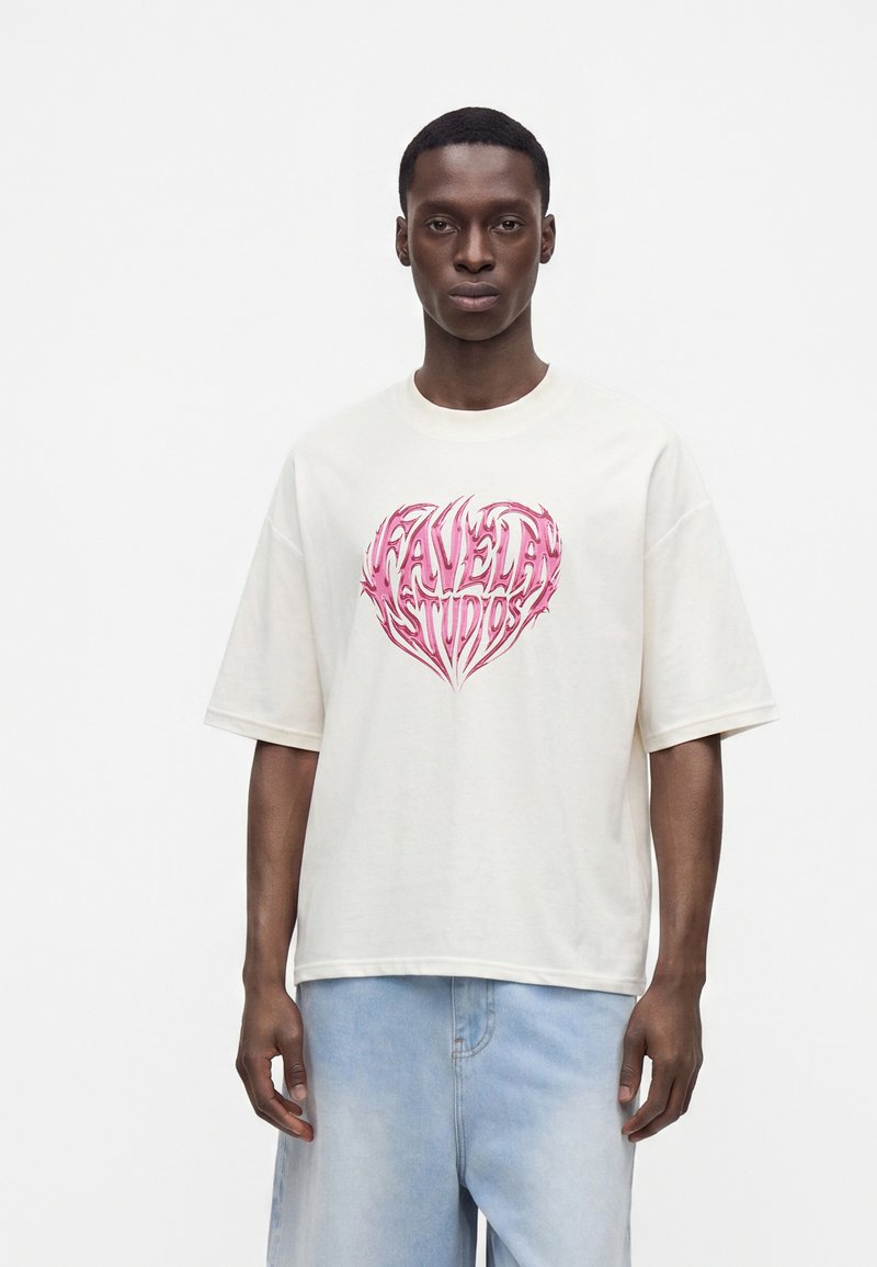 Νεαρός άνδρας φοράει λευκό oversized μπλουζάκι με ροζ καρδιά και ανοιχτό μπλε τζιν, στέκεται μπροστά σε απλό λευκό φόντο.