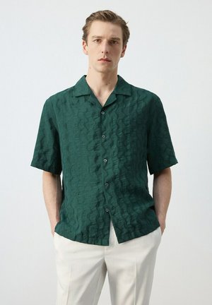 Joven con camisa de manga corta abotonada texturizada en verde oscuro y pantalones beige claros, de pie con las manos en los bolsillos.