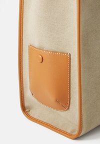 Sac en toile beige avec des accents en cuir marron, présentant une poche à rabat avec un bouton-pression et des coutures visibles le long des bords.