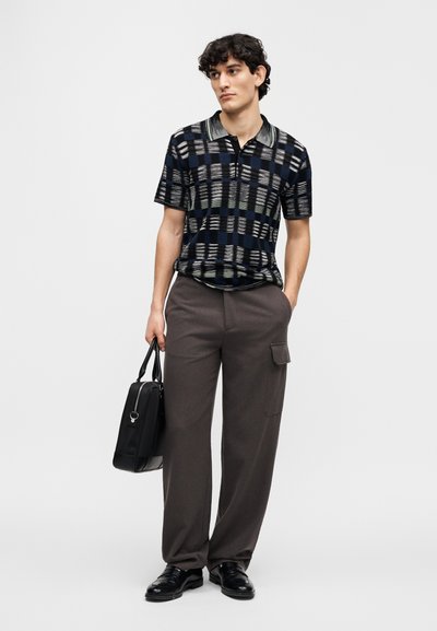 Short-sleeve, black and blue patterned polo shirt with a collar, paired with gray high-waisted trousers and black shoes, carrying a black bag.