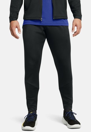 Schwarze Sportsleggings mit einem schmal zulaufenden Design, glatter Textur und kantigen Einsätzen. Kombiniert mit schwarzen Sneakers, die blaue Schnürsenkel haben.
