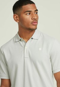 Polo de color gris claro con un acabado texturizado, de manga corta y con un cuello clásico, que presenta un pequeño logo en el lado izquierdo del pecho.
