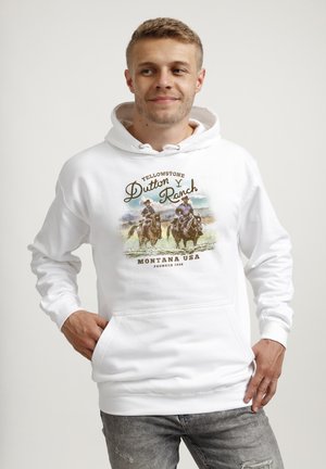 Młody mężczyzna w białej bluzie z kapturem z grafiką przedstawiającą dwóch kowbojów na koniach oraz tekstem "Yellowstone Dutton Ranch Montana USA Founded 1886."