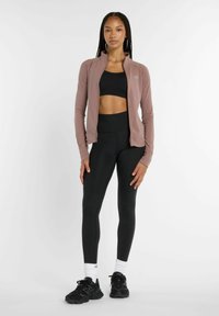 Giacca zip a luce rosa sopra un crop top nero e leggings neri a vita alta. Sneakers nere e calzini bianchi alla caviglia completano l'outfit.