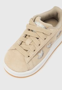 Μπεζ σουέτ αθλητικό παπούτσι με λευκή λαστιχένια σόλα, στρογγυλή μύτη, υφασμένα κορδόνια και διακοσμημένο με διαφανή στοιχεία στις πλευρές.