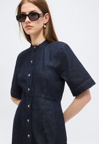 Γυναίκα με σκουρόχρωμο φόρεμα από τζιν με κουμπιά και κοντά μανίκια, μαύρα γυαλιά ηλίου και μεγάλα ασημένια σκουλαρίκια με κρεμαστά στοιχεία, μπροστά σε ένα απλό ανοιχτόχρωμο φόντο.