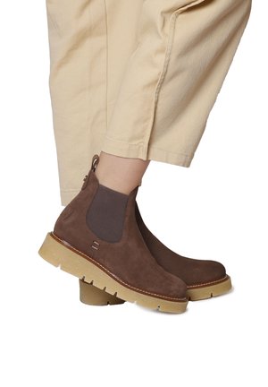 Stivaletti in suede marrone con pannelli elastici laterali, suola beige chunky e cuciture marroni, abbinati a pantaloni tapered beige chiaro.