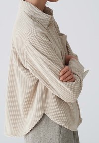 Chemise en velours côtelé beige avec des côtes verticales, un col classique et une fermeture à boutons. Dotée d'un ourlet arrondi et de manches larges.