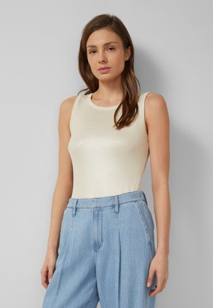 Vrouw met lang bruin haar die een mouwloze witte top en lichtblauwe jeans met hoge taille draagt, staand tegen een grijze achtergrond.