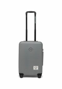 Valise rigide grise avec une surface lisse, poignée noire rétractable, quatre roues et une étiquette Herschel blanche à l'avant.