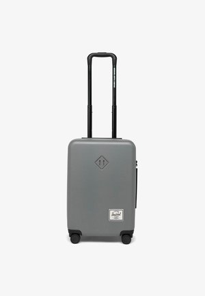 Valise rigide grise avec une surface lisse, poignée noire rétractable, quatre roues et une étiquette Herschel blanche à l'avant.