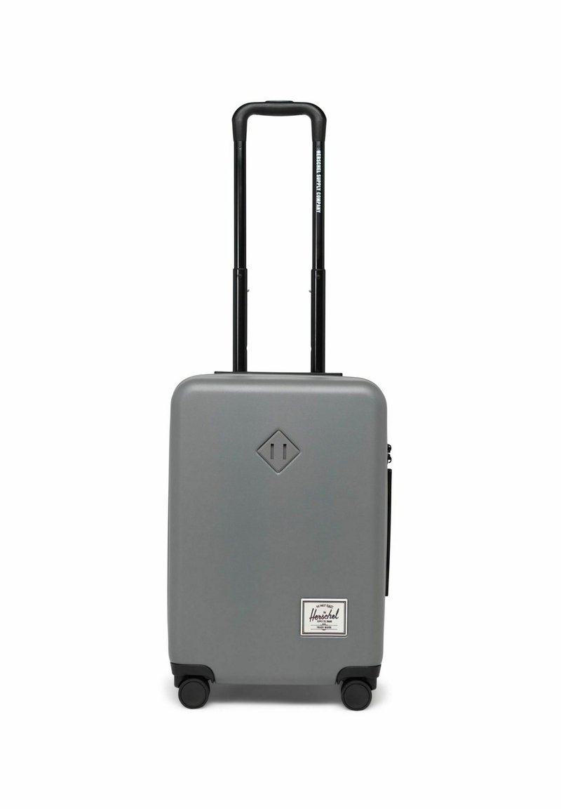 Valise rigide grise avec une surface lisse, poignée noire rétractable, quatre roues et une étiquette Herschel blanche à l'avant.