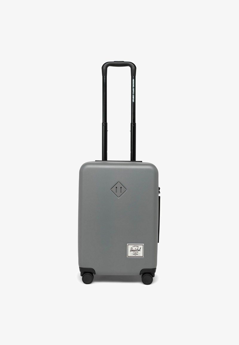 Valise rigide grise avec une surface lisse, poignée noire rétractable, quatre roues et une étiquette Herschel blanche à l'avant.