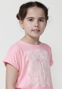 Rožinė trumpomis rankovėmis marškinėliai su sudėtingu baltu siuvinėjimu, vaizduojančiu gėlių ir plunksnų raštus. Minkšta medžiaga su laisvu krovu.