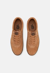 Sapatilhas castanhas de camurça com um acabamento texturizado, design com atacadores e logótipo preto da Vans na língua. Sola em borracha visível nas bordas.