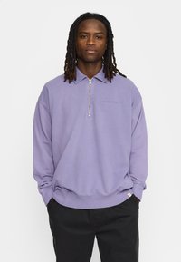 Lichtpaarse zip-up sweatshirt met een kraag, voorzien van een subtiele reliëflogo. Zachte textuur en een losse pasvorm. Gecombineerd met zwarte broeken.