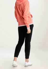 Korallfärgad hoodie med snörning och vita detaljer, parat med svarta joggerbyxor och vita träningsskor. Slät tygkvalitet och avslappnad passform.