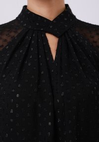 Blouse noire en tissu transparent avec motif à pois texturé, col en trou de serrure et design plissé. Texture lisse avec une brillance subtile.