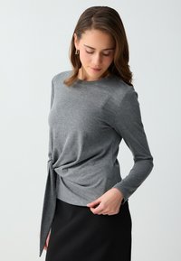 Femme portant un haut gris à manches longues avec un nœud sur le côté et une jupe noire, regardant vers le bas à sa gauche sur un fond blanc.