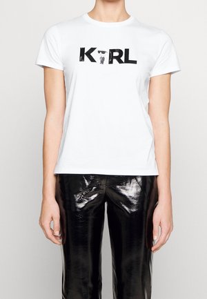 Person, die ein weißes T-Shirt mit dem Text "KARL" und einer Cartoonfigur zwischen den Buchstaben trägt, kombiniert mit glänzenden schwarzen Hosen vor weißem Hintergrund.
