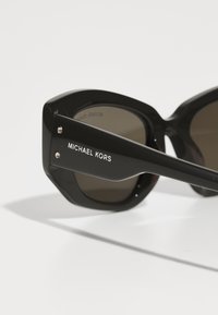 Černé oversized sluneční brýle s geometrickým rámem, gradientovými skly a stříbrným logem "MICHAEL KORS" na spáncích.