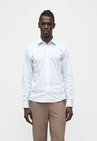 Homme debout portant une chemise blanche à boutons avec des motifs floraux bleu clair et un pantalon marron clair sur un fond neutre.