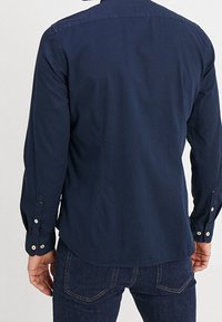 Mann in einem langärmeligen marineblauen Hemd und dunkelblauen Jeans, von hinten gezeigt, vor einem einfarbigen hellen Hintergrund stehend.