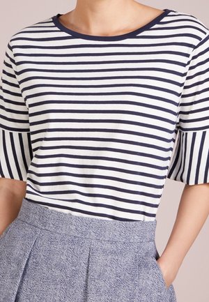 Frau trägt ein kurzärmeliges Oberteil mit horizontalen Marineblau- und Weißstreifen, das in einen hoch taillierten, strukturierten blaugrauen Rock gesteckt ist.