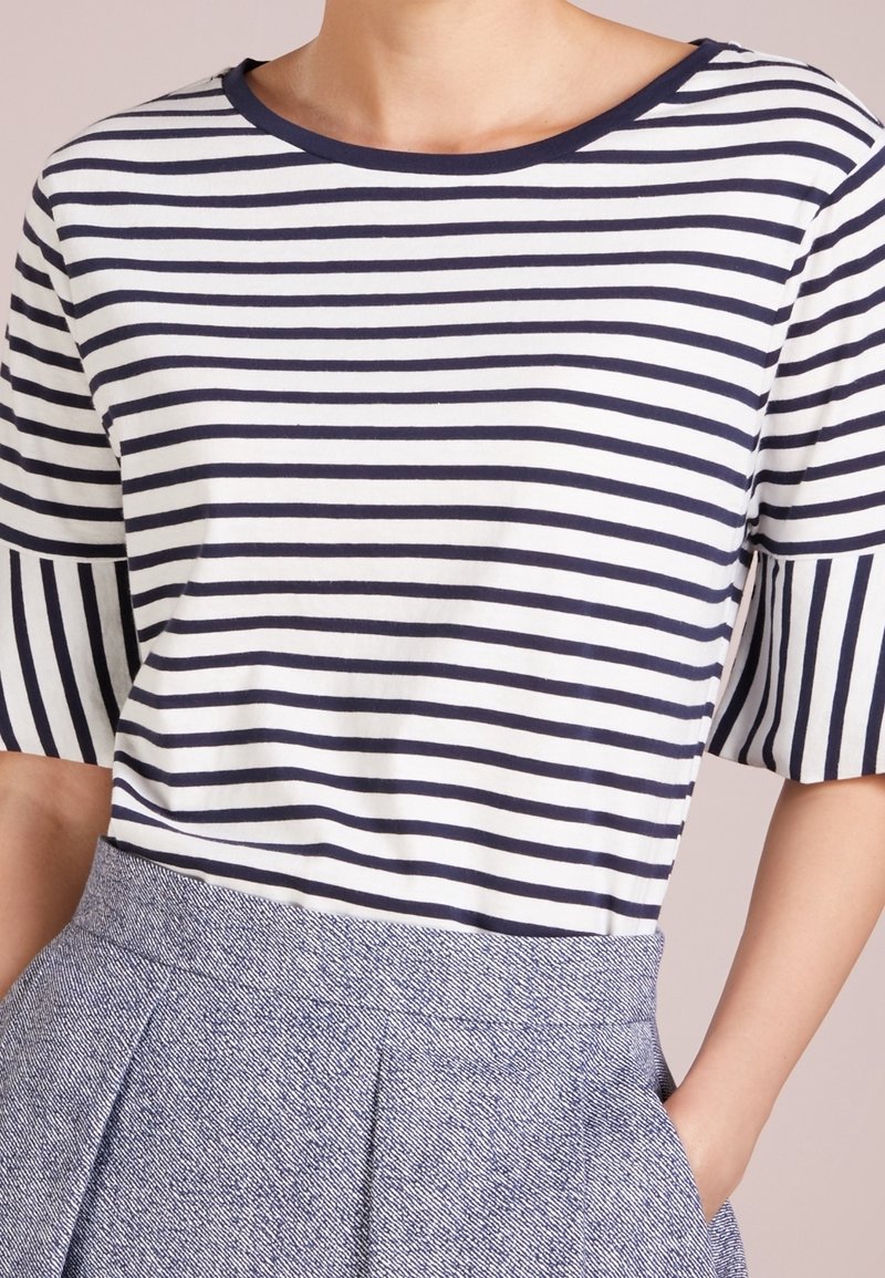 Kobieta ubrana w granatowo-białą, poziomo paskowaną koszulkę z krótkim rękawem, wpuszczoną w wysokowęzią spódnicę o fakturze w kolorze niebiesko-szarym.
