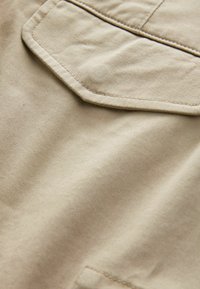 Beige puuvillakangas, jossa on sileä rakenne, sisältää ommellun taskuyksityiskohdan. Materiaali näyttää hienoa nauhakuviota ja siistiä viimeistelyä.