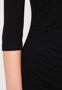 Robe noire ajustée à manches trois-quarts, avec des plis texturés et un tissu doux et extensible, mettant en valeur une silhouette élégante.