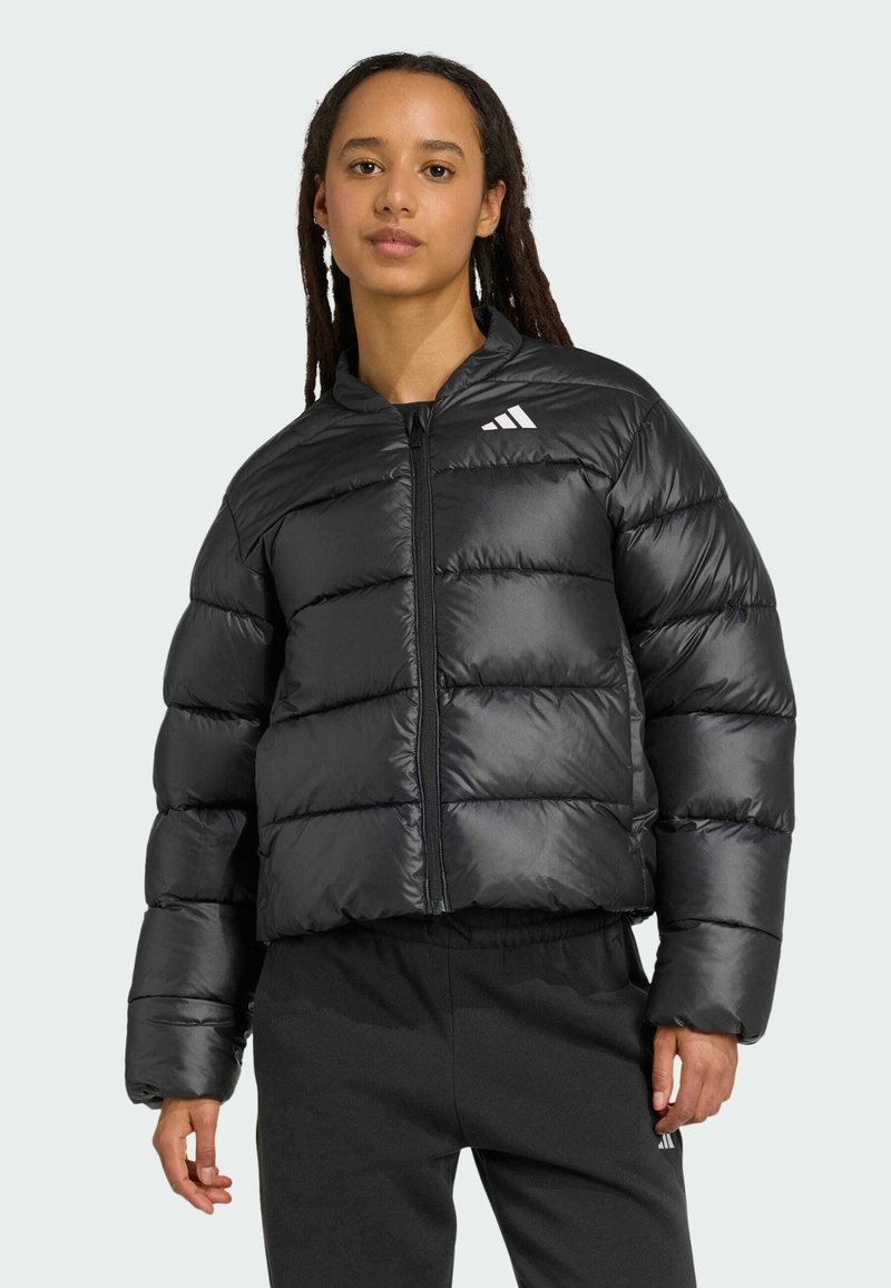 Mladý človek s dredmi má na sebe čiernu puffrovú bundu a tmavé nohavice, stojí pred svetlo šedým pozadím.