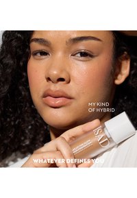 A clear glass bottle of foundation with a white cap. The label displays "Wake Up The Glow" and contains a liquid with a luminous finish.