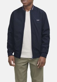 Giacca bomber navy con colletto a costine, zip frontale e polsini elasticizzati, indossata sopra un maglione chiaro e abbinata a pantaloni beige.