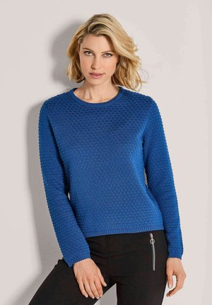 Blonde Frau in einem strukturierten blauen Pullover und schwarzen Hosen mit einem silbernen Reißverschlussdetail, die vor einer hellen Wand steht.