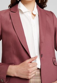 Burgundské přiléhavé sako s hladkou teksturou, zapínání na jeden knoflík a klopové límce. V kombinaci s bílou košilí s knoflíky a vzorovanými kalhotami.