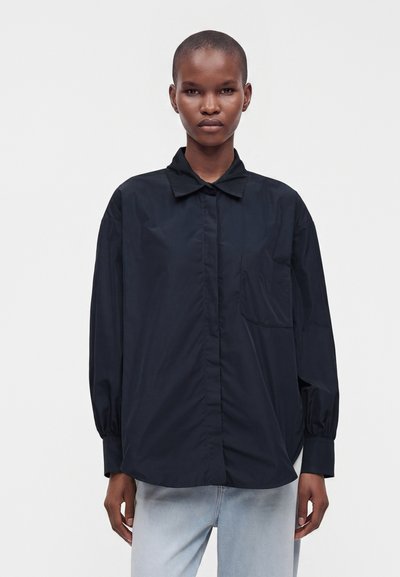 Personne portant une chemise noire ample à manches longues avec col et poche avant, associée à un jean bleu clair sur un fond uni.