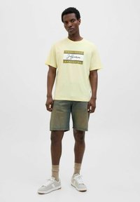 Gul bomulds t-shirt med et trykt logo-design på forsiden, parret med slidte denim shorts. Fodtøj: grå og hvide sneakers med beige sokker.