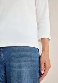 Wit shirt met driekwartmouwen gecombineerd met blauwe denim jeans. Het shirt heeft een gladde textuur, terwijl de jeans een licht versleten afwerking hebben.
