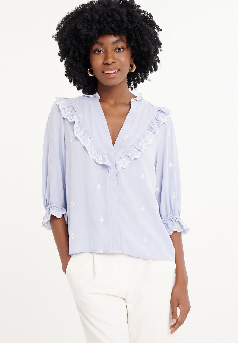 Blusa azzurro chiaro con colletto e polsini con volant, caratterizzata da motivi ricamati bianchi. Vestibilità rilassata con maniche a tre quarti e scollatura a V.