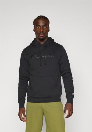 Hanorac negru din bumbac, cu un buzunar frontal, șnururi și un logo brodat "Champion" pe piept. Purtați cu pantaloni de culoare verde măsliniu.