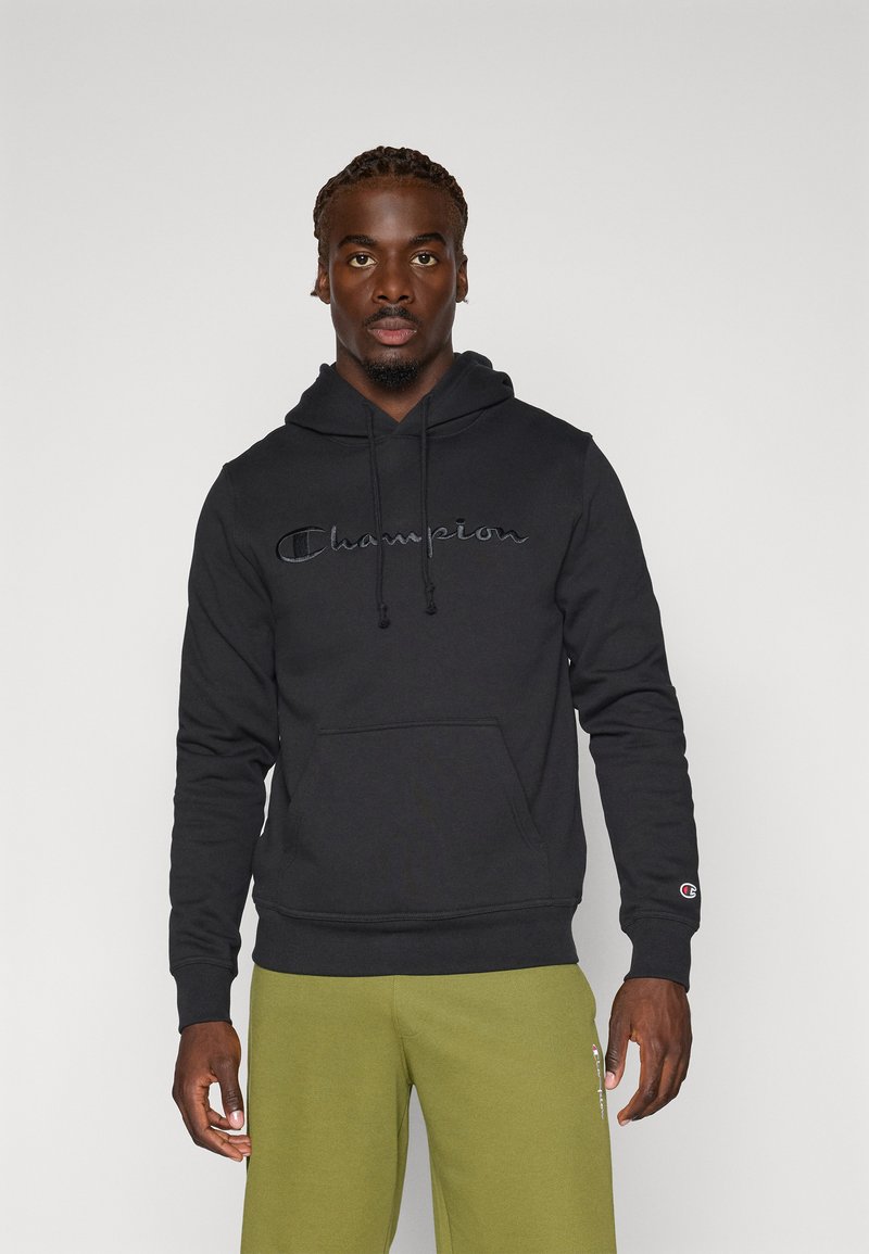 Hoodie noir en coton, avec une poche avant, des cordons de serrage et un logo "Champion" brodé sur la poitrine. Porté avec un pantalon vert olive.