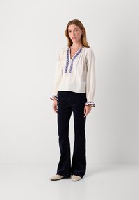 Weiße bestickte Bluse mit blauen Mustern, gerafften Ärmeln und Rüschensäumen, kombiniert mit dunkelblauen ausgestellten Cordhosen und beigefarbenen Schuhen.