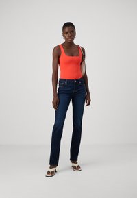 Φωτεινή πορτοκαλί ολόσωμη φόρμα με τετράγωνη λαιμόκοψη, συνδυασμένη με σκούρα μπλε τζιν με ίσια γραμμή. Η μοντέλο φοράει ανοιχτόχρωμα τακούνια και κρατάει μια τσάντα.