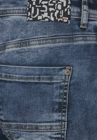 Jeans de mezclilla en azul oscuro, con una tela texturizada, costuras amarillas en contraste y un parche negro con patrones abstractos en blanco.