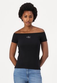 Juodas off-the-shoulder marškinėliai, pagaminti iš minkštos medžiagos, su mažą baltą Calvin Klein logotipą ant krūtinės ir trumpomis rankovėmis.