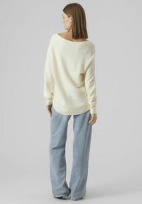 Hellgelb gestrickter Pullover mit überschnittenen Schultern, weitem Ausschnitt und langen Ärmeln. Kombiniert mit lockeren blauen Jeans, von hinten betrachtet.