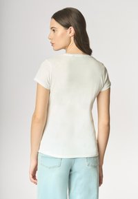 T-shirt bianco a maniche corte con scollo rotondo, texture liscia e dettaglio a bottone sul retro, abbinato a jeans azzurri chiari.