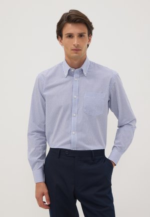 Giovane uomo che indossa una camicia a quadri azzurri chiari con maniche lunghe e pantaloni blu scuro, in piedi davanti a uno sfondo semplice.