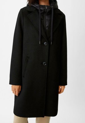 Femme portant un manteau en laine noir à deux boutons sur une veste noire à capuche zippée avec cordons et un pantalon beige.